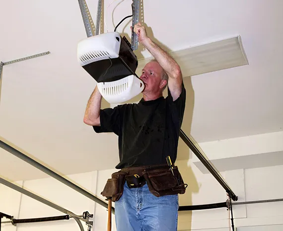 Technician inspecting a garage door opener mechanism while standing on a ladder.