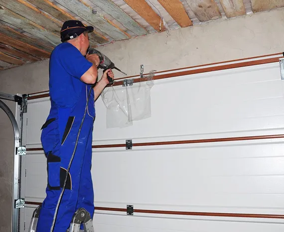 A technician wearing blue overalls working on a garage door spring mechanism inside a garage.