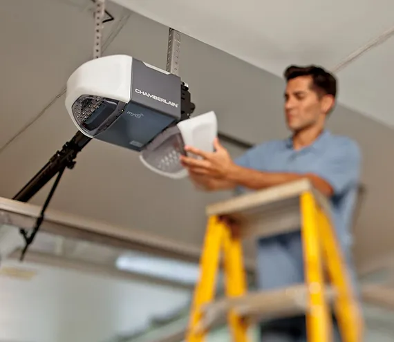 Technician on a yellow ladder installing a garage door opener inside a garage.